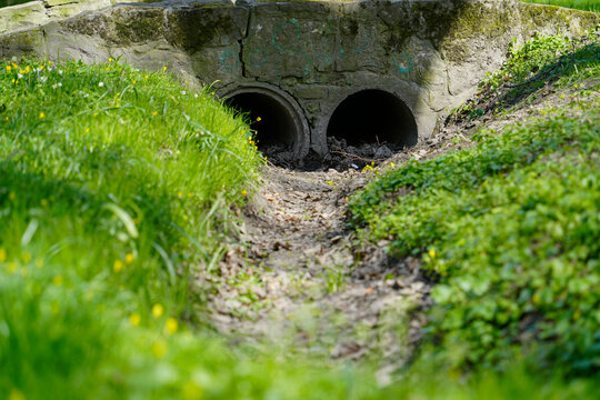 Concrete culvert outlet with two drainage pipes in overgrown grassy ditch. Functional infrastructure visual ideal for civil engineering or water runoff topics