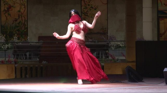 A belly dancer in a red costume performs on stage. She wears a matching face covering. The stage is lit with colorful lights, and there are decorations in the background.