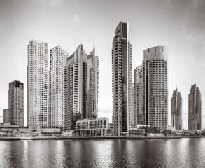 Monochrome city skyline with tall skyscrapers reflecting on calm water under a cloudy sky, Dubai Marina bay UAE