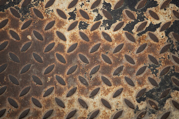 Close-up view of a rusty metal surface featuring a diamond pattern. The texture exhibits wear and aging effects, revealing industrial character and unique design qualities over time