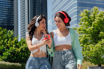 Two young women sharing earphones listening to music on a smartphone in the city