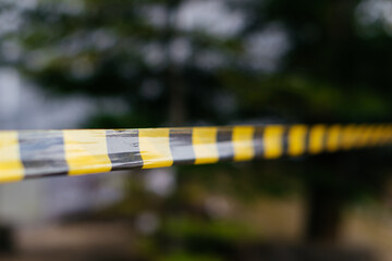 A close-up of a yellow and black caution tape stretched across an outdoor scene, symbolizing...
