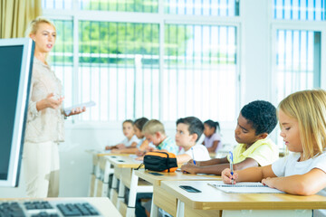 Focused preteen pupils studying in classroom with female teacher, writing exercises in workbooks