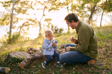 Fototapeta premium Laughing dad with a little daughter are sitting on stumps in a sunny meadow in the forest