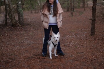 A woman wearing a beige puffer jacket and blue jeans stands in a forest, holding her white dog on a...