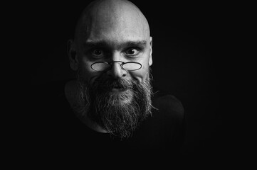 Black and white portrait of a tense bald man with a gray beard and round glasses makes a slight...