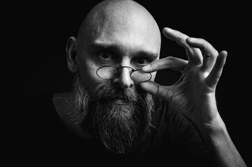 Black and white portrait of a tense bald man with a gray beard and round glasses makes a slight pinching gesture, emotions. Low key portrait with dramatic lighting and expressive face on a black backg