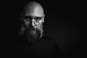 Black and white portrait of a tense bald man with a gray beard and round glasses makes a slight...