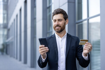 Fototapeta premium A businessman looks at his phone while holding a credit card. He appears to be making a payment with mobile banking or online.