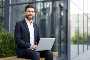 A smiling businessman sits outside, working on a laptop. He wears a suit and is positioned in front of a modern building, suggesting a professional setting.
