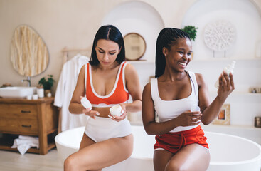 Happy diverse friends enjoying personal grooming moment in modern bathroom space. Cheerful multiracial Women in sportswear using cosmetics and cream while performing mindful skin assessment