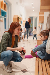 Preschool teacher helps a young girl with her shoes in the hallway, showing care and attention...