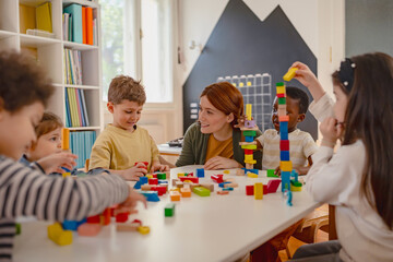 Fototapeta premium Preschool teacher interacts with a diverse group of children building with colorful blocks in a bright classroom, promoting creativity, teamwork, and multicultural early education.
