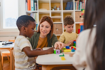 Preschool teacher interacts with a diverse group of children building with colorful blocks in a...