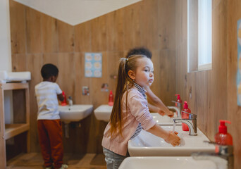 Multicultural preschool children wash their hands at sinks in a wooden-themed bathroom, practicing hygiene routines as part of their daily classroom activities.