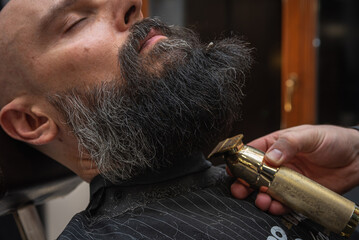 Close-up of a barber trimming a beard with electric clippers in a modern barbershop. Professional men's grooming and beard care service, selective focus