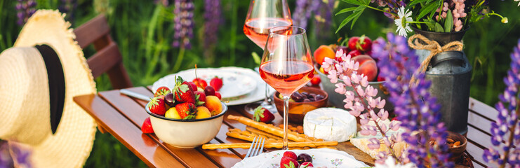 Banner. Elegant gorgeous wedding table decor or romantic dinner arrangement outdoors in blooming field. Purple lupine flowers, candles, fruits, wine, wooden furniture. Sunset, summer, golden hour