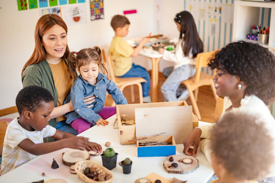 Diverse group of preschool children engage in a nature-based learning activity with two teachers, using wood and natural objects to explore creativity and tactile skills in the classroom.