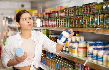 Fototapeta premium Young woman looking to buy jar of mayonnaise in supermarket