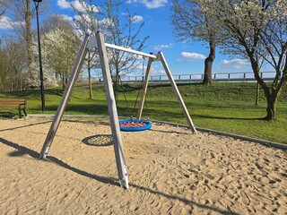A round basket swing stands on a sandy playground in Smardzewice, surrounded by green grass and blooming trees under a bright blue sky.