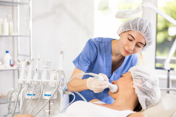 Naklejka premium Positive young female cosmetologist performing hardware facial procedure on woman using high frequency ultrasonic vibration attachment in cosmetic clinic to improve efficacy of skincare products