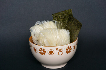 Two portions of wide raw rice noodles with two nori sheets in a soup bowl on a black background. Asian cuisine preparation. Close-up.