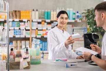 Young professional female parmacist working computer and helping young guy with prescription in hand look for medicine in big drugstore