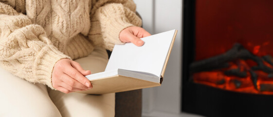 Young woman reading book in armchair near fireplace at home, closeup
