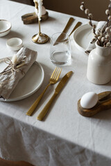 Spring minimalist table setting in warm neutral colors. Easter festive table decoration, white plates, gold cutlery, candles, eggs and willow branches in vase