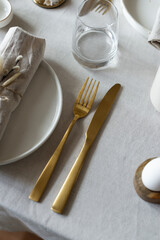 Spring minimalist table setting in warm neutral colors. Easter festive table decoration, white plates, gold cutlery, candles, eggs and willow branches in vase