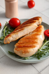 Plate of tasty grilled chicken breast with arugula and tomatoes on white tile background