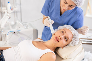 Young woman cosmetologist performs hardware facial microcurrent procedure to young female patient