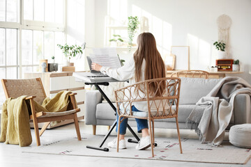 Young woman with note sheets playing synthesizer in living room, back view