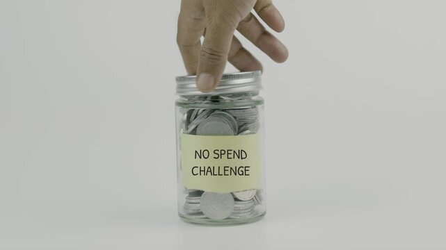No Spend Challenge text or word on yellow sticky notes attached to a coin jar, hand inserting a coin before sealing the lid, symbolizing the commitment to saving money by avoiding unnecessary spending