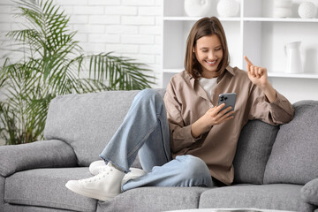 Obraz premium Young happy woman holding mobile phone with smart home security system control panel on sofa in room