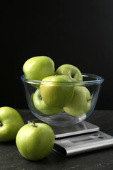 Electronic kitchen scale with bowl of green apples on dark textured table against black background, closeup