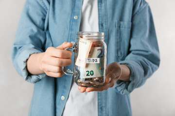 Woman holding tips jar with money on light background, closeup