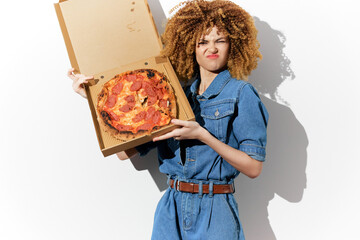 Young woman with curly hair holding a pizza box, expressing playful disgust in a denim outfit...