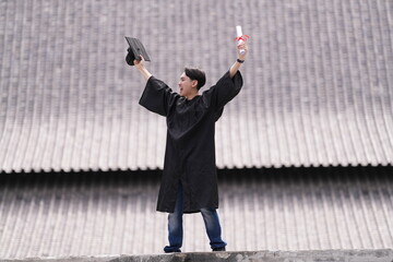 A proud graduate in a black gown holds his cap and diploma high in celebration, smiling against a...