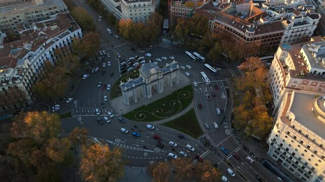 Stunning aerial drone footage showcasing the iconic Puerta de Alcal in Madrid, beautifully lit by the sunset, surrounded by vibrant autumn foliage and car traffic