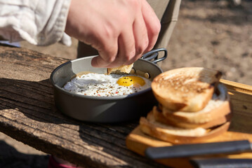 Unknown traveler eating breakfast outdoors, fried eggs and toasted bread slices. Close up composition with selective focus on the piece of bread to be dipped in the egg yolk.