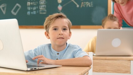 Attractive caucasian boy looking at camera while studying math at school. Diverse cute student doing test while teacher asking asian child question and point at paper in front of blackboard. Pedagogy.