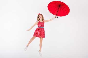 Young woman in retro polka dot dress posing with red umbrella against white background, capturing...
