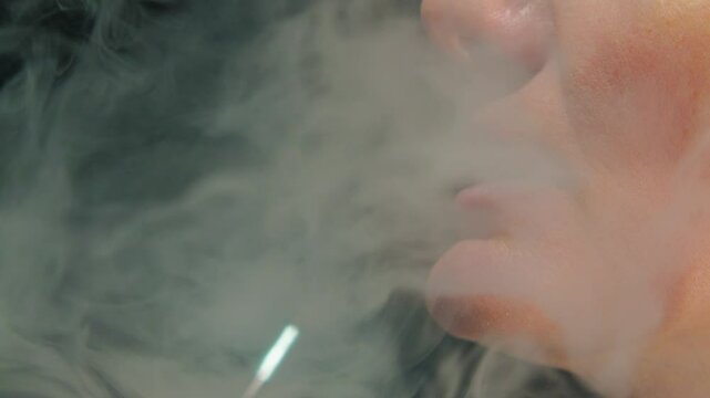 Mouth of caucasian woman exhaling thick vape smoke on black background. Close-up of electronic cigarette smoking. Side view