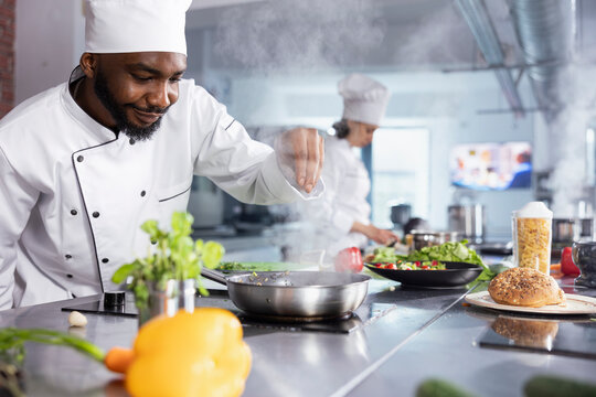 In a kitchen, black chef adding fresh herbs to complete a gourmet dish, balancing flavors with rosemary and other condiments. Kitchen counter stocked with vegetables and food prep essentials.