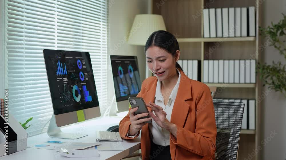 Poster young businesswoman using smartphone while sitting at desk in modern office with computer and docume