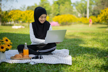 Young Asian Muslim female student sitting on mat, using notebook at summer garden park. Isalamic business woman enjoy relaxing with laptop and picnic at public.