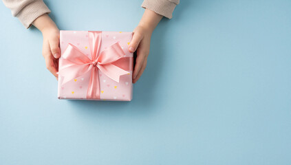 A pair of hands presents a beautiful pink gift box adorned with a ribbon, set against a soft blue...