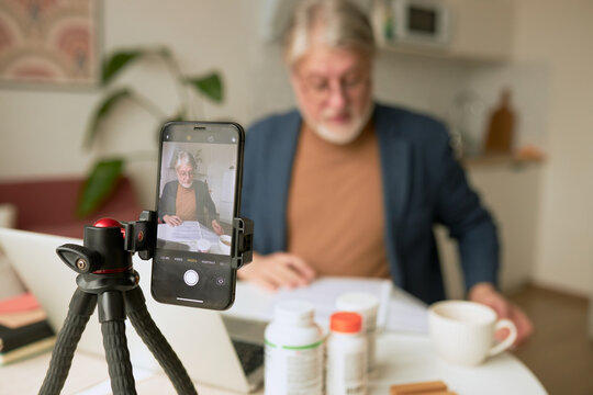 Selective focus on phone mounted on tripod recording video of senior qualified male nutritionist of 60s surrounded with bottles of food supplement, reading paper doc, giving advice to stay healthy