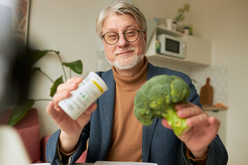 Senior man in glasses holding bottle of food supplements and green head of broccoli, choosing...